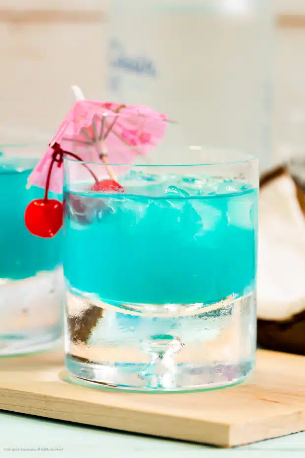 Straight on photo of a blue hawaiian cocktail garnished with maraschino cherries and a cocktail umbrella.