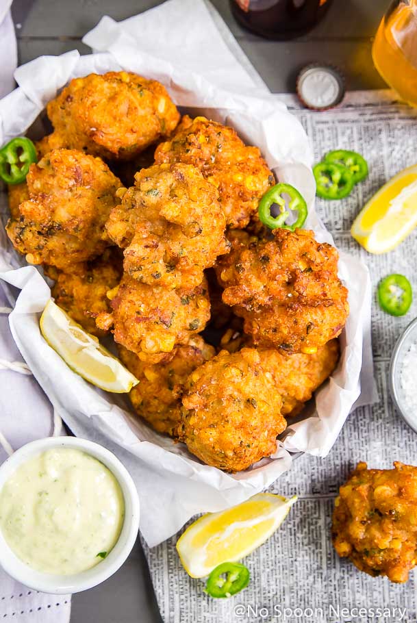 Overhead shot of a basket lined with white paper and filled with Lobster and Bacon Corn Fritters; with a small bowl of jalapeno-honey aioli, lemon wedges, jalapeno slices, beer bottle, beer cap and newspaper scattered in the background
