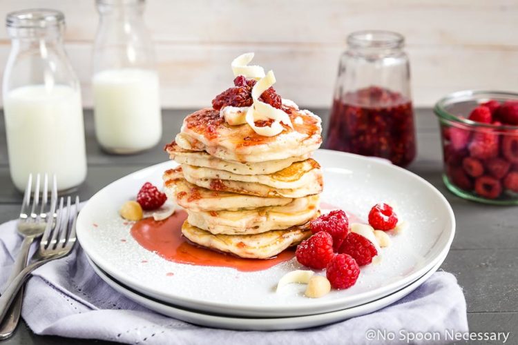 Straight on shot of a stack of macadamia nut pancakes topped with raspberry syrup and white chocolate shavings; with a glass jar of milk, bowl of raspberries, forks and glass jar of syrup in the background.