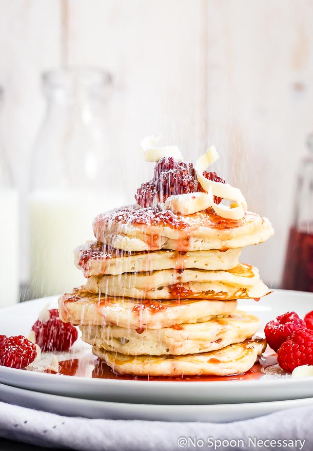 Straight on shot of powdered sugar being sprinkled on a stack of macadamia nut pancakes topped with raspberry syrup and white chocolate shavings; with glass jars of milk blurred in the background.