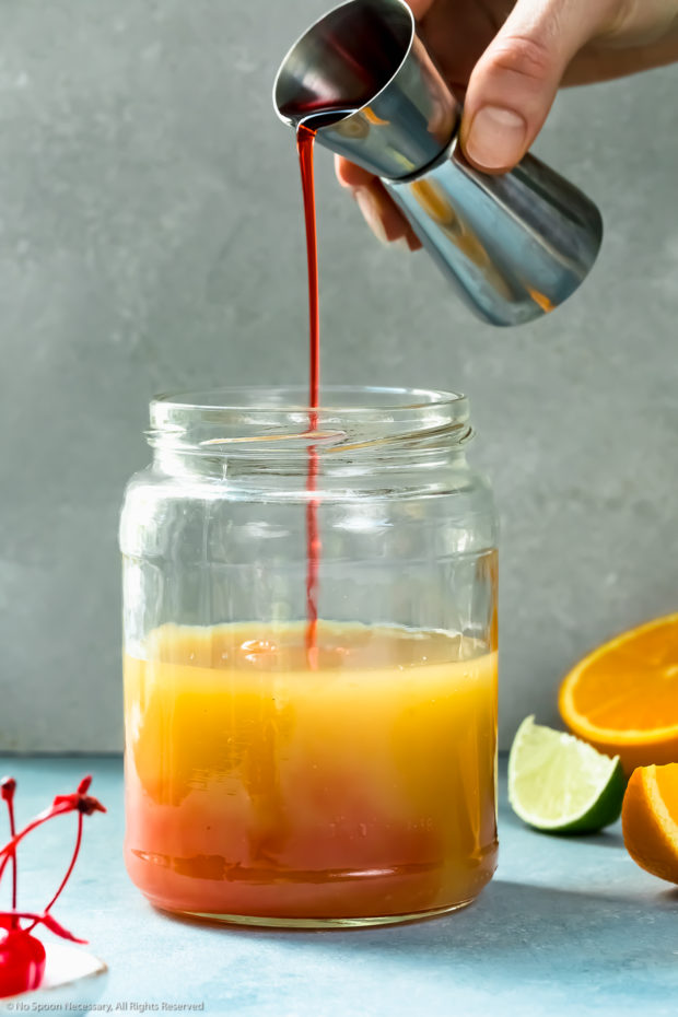 Straight on photo of a mason jar filled with rum, liqueur and fruit juices with a shot of grenadine being poured into the jar - photo of how to make a rum runner cocktail.