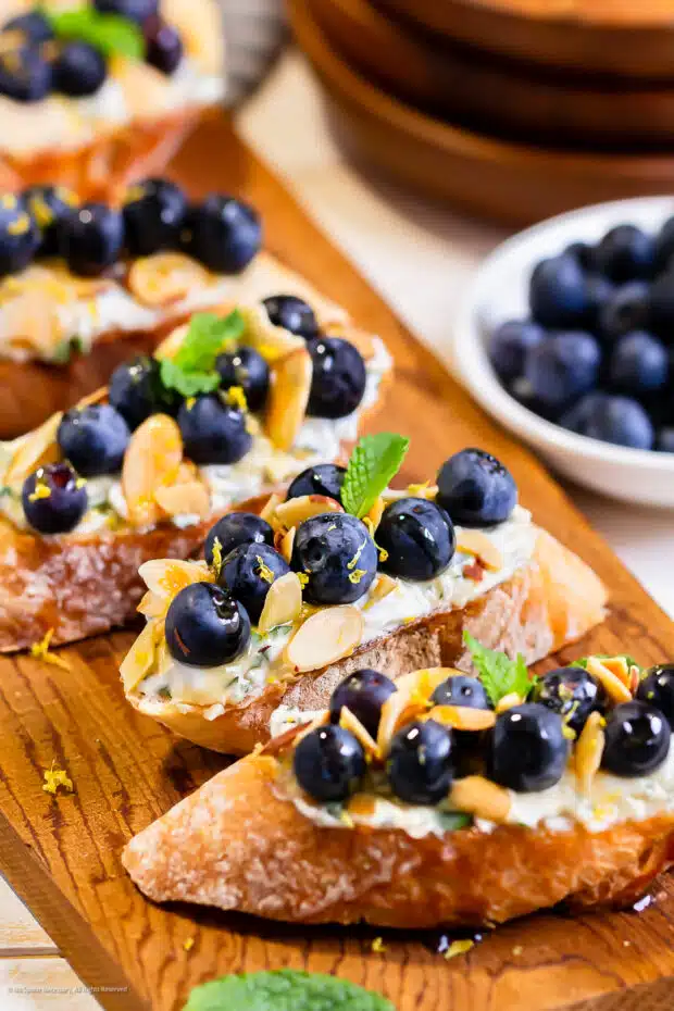 Angled photo of four toasted slices of crostini topped with fresh blueberries and mint whipped ricotta cheese.
