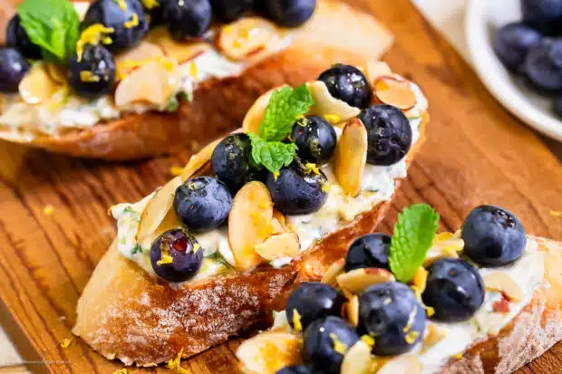 Angled photo of a slice of crispy bread topped with blueberries, whipped ricotta cheese, almonds, and honey.