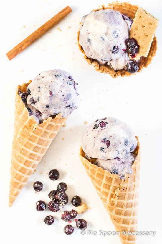 Overhead shot of 3 scoops of No Churn Blueberry Cinnamon Ice Cream in a waffle cones with fresh blueberry compote and a cinnamon stick next to the cones.