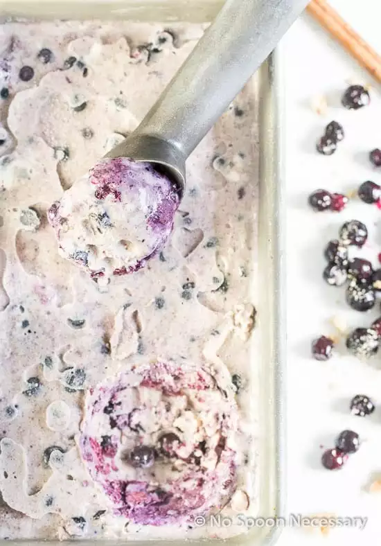 Overhead shot of No Churn Blueberry Cinnamon Ice Cream in a silver container with an ice cream scoop and fresh blueberry compote and a cinnamon stick scattered next to the container.