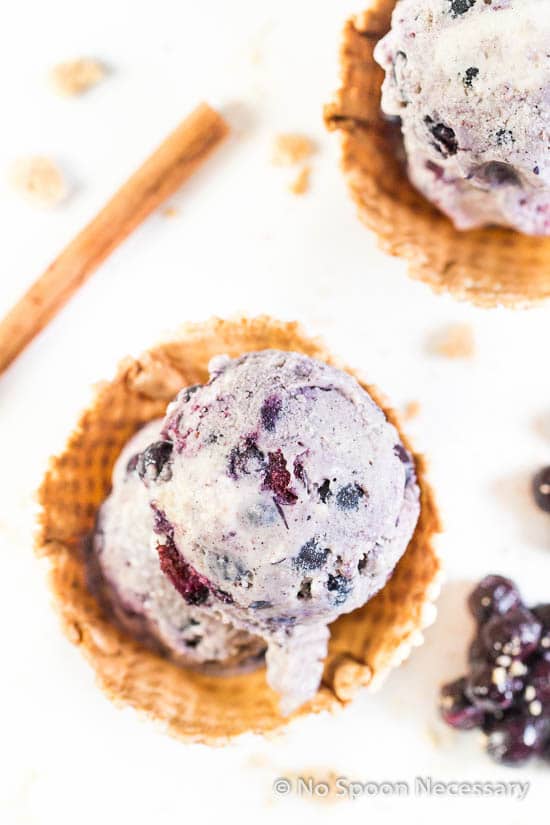 Overhead shot of 2 scoops of No Churn Blueberry Cinnamon Ice Cream in a waffle bowls with fresh blueberry compote and a cinnamon stick scattered around the bowls.