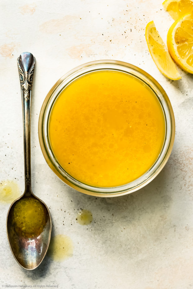 Overhead photo of lemon vinaigrette for couscous in a glass jar with a mixing spoon and fresh lemon wedges next to the jar.