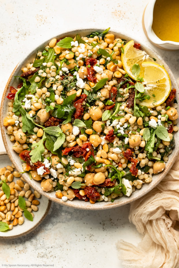 Overhead photo of Israeli couscous with sun-dried tomatoes, arugula and chickpeas in a large white bowl with a jar of lemon vinaigrette and ramekin of toasted pine nuts next to the bowl.
