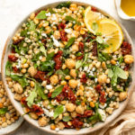 Overhead landscape photo of Israeli pearl couscous with sun-dried tomatoes, arugula and chickpeas in a large white bowl with a jar of lemon vinaigrette and ramekin of toasted pine nuts next to the bowl.