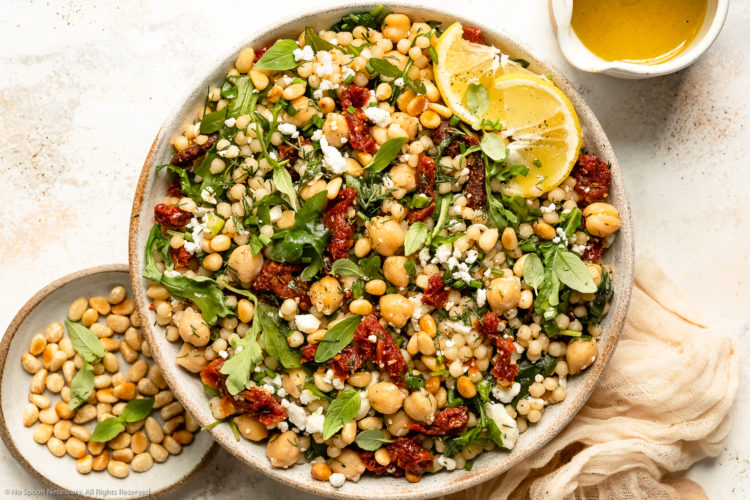 Overhead landscape photo of Israeli pearl couscous with sun-dried tomatoes, arugula and chickpeas in a large white bowl with a jar of lemon vinaigrette and ramekin of toasted pine nuts next to the bowl.