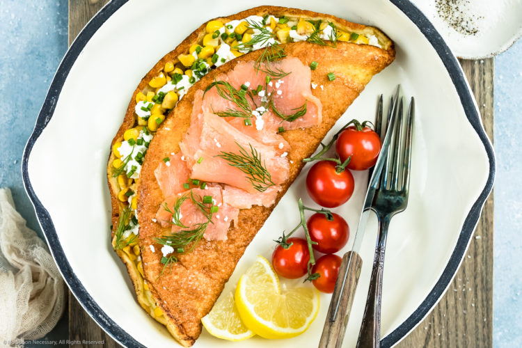 Overhead, landscape shot of a fluffy omelet filled with corn, goat cheese and chives in a white skillet with cherry tomatoes, lemon wedges and a knife and fork next to the omelet.