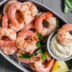 Overhead shot of a small gray tub filled with ice and topped with Perfect Beer Poached Shrimp Cocktail with Remoulade in a small ramekin with a shrimp laying on the sauce; with fresh parsley and shrimp forks on either side of the tub.