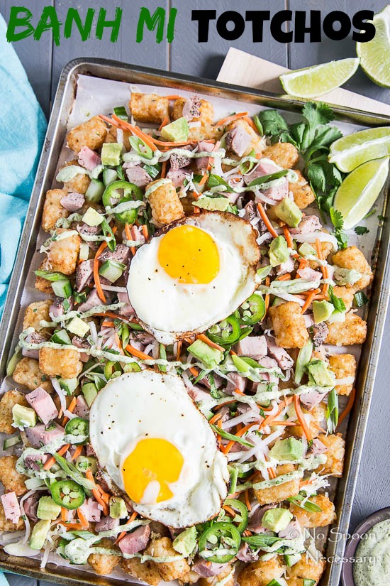 Overhead shot of Banh Mi Totchos (tater tot nachos) on a sheet pan topped with two fried eggs; with lime wedges and a ramekin of cilantro lime aioli in the right corners of shot.