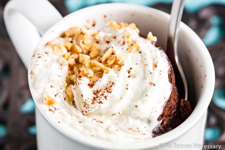 Angled, landscape shot of a Snickers Lava Mug Cake topped with whipped cream and crushed peanuts in a white mug with a spoon inserted into the mug.
