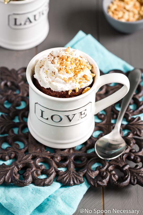Angled shot of a Snickers Lava Mug Cake topped with whipped cream and crushed peanuts in a white mug with a spoon and teal linen next to it