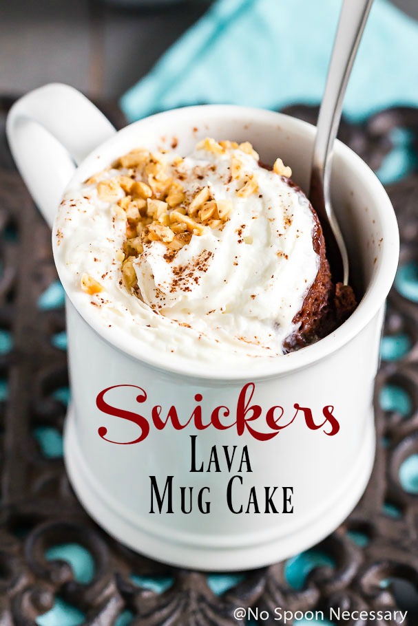Angled shot of a Snickers Lava Mug Cake topped with whipped cream and crushed peanuts in a white mug with a spoon inserted into the mug.