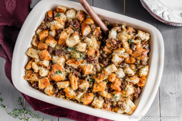Overhead shot of Caramelized Onion, Sausage, Pecan & Croissant Stuffing in a baking dish with fresh thyme and a stack of plates surrounding the dish.