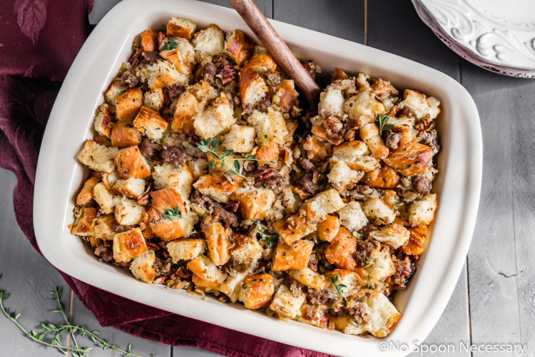 Overhead shot of Caramelized Onion, Sausage, Pecan & Croissant Stuffing in a baking dish with fresh thyme and a stack of plates surrounding the dish.