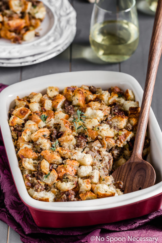 Angled shot of Caramelized Onion, Sausage, Pecan & Croissant Stuffing in a baking dish with a serving spoon inserted into the dish and a plate of stuffing and wine glass blurred behind the dish.