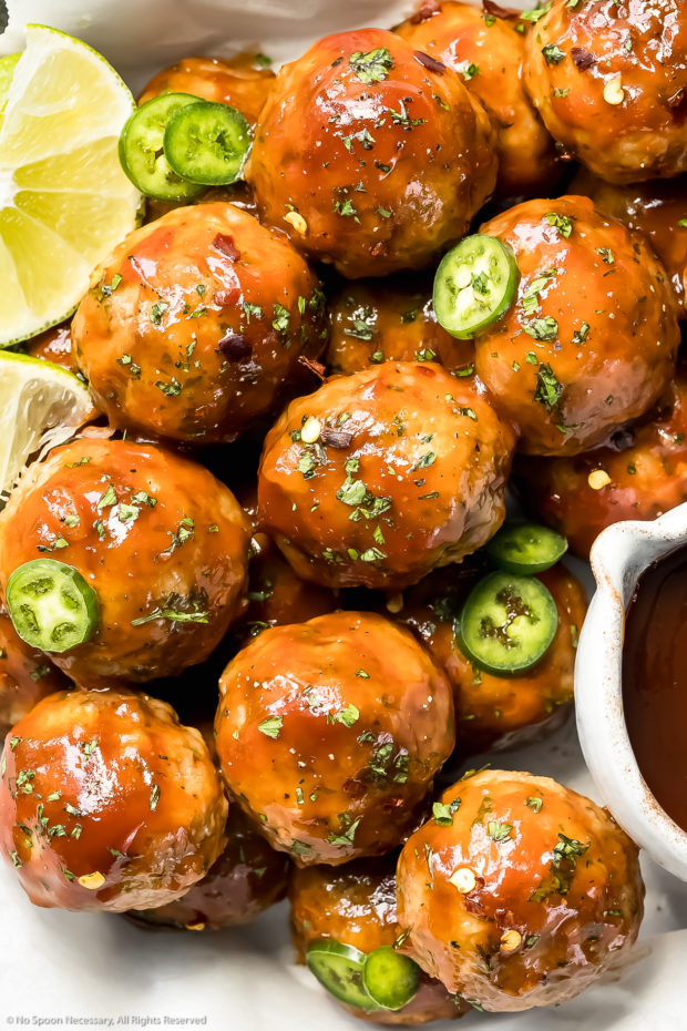 Overhead up-close photo of Dr Pepper Meatballs garnished with chopped parsley and sliced jalapenos with lime wedges and a ramekin of dr pepper glaze next to the meatballs.