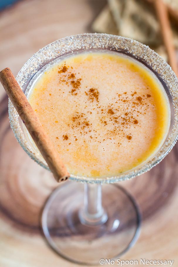 Slightly angled shot of a Pumpkin Pie Martini garnished with a cinnamon stick and graham cracker-sugar rim with cinnamon sticks and graham crackers blurred in the background.