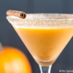 Straight on landscapt shot of a Pumpkin Pie Martini garnished with a cinnamon stick and graham cracker-sugar rim with a small pumpkin blurred in the background.