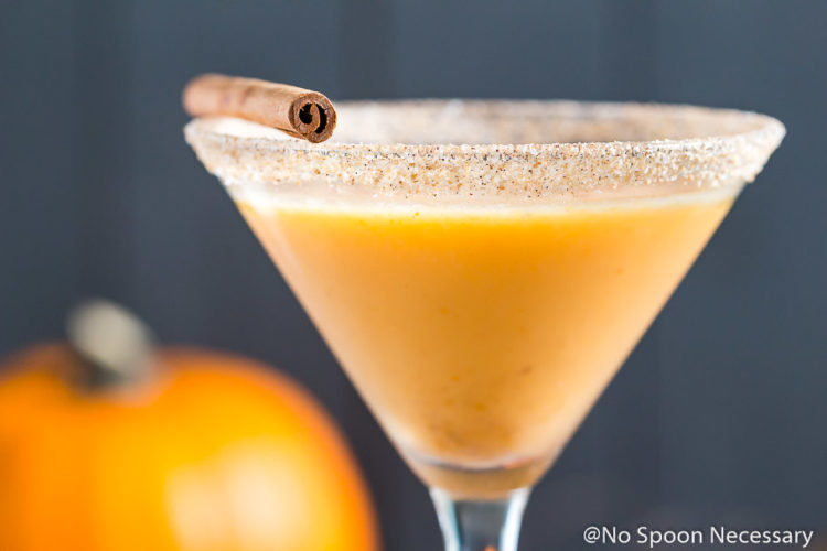 Straight on landscapt shot of a Pumpkin Pie Martini garnished with a cinnamon stick and graham cracker-sugar rim with a small pumpkin blurred in the background.