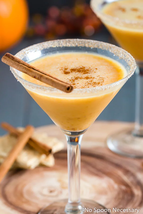 Angled shot of a Pumpkin Pie Martini garnished with a cinnamon stick and graham cracker-sugar rim with cinnamon sticks, graham crackers, a small pumpkin and autumn decor blurred in the background.