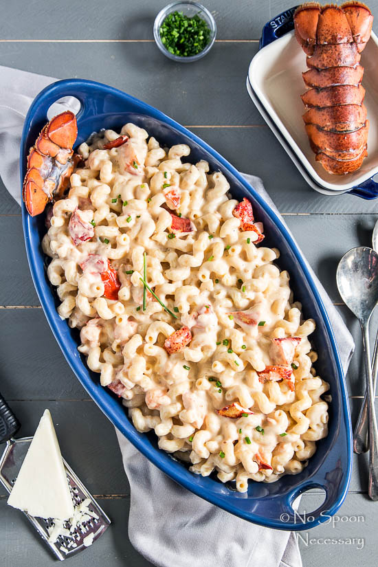 Overhead shot of Truffle Lobster Macaroni n' Cheese in a large blue baking dish on a gray linen with a chunk of parmesan on a grater in the bottom corner and a ramekin of chives, lobster tail and serving spoons in the upper corners.