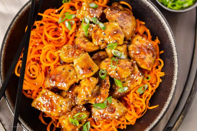 Overhead photo of stir fried honey garlic chicken breast with carrot noodles in a large black serving bowl.
