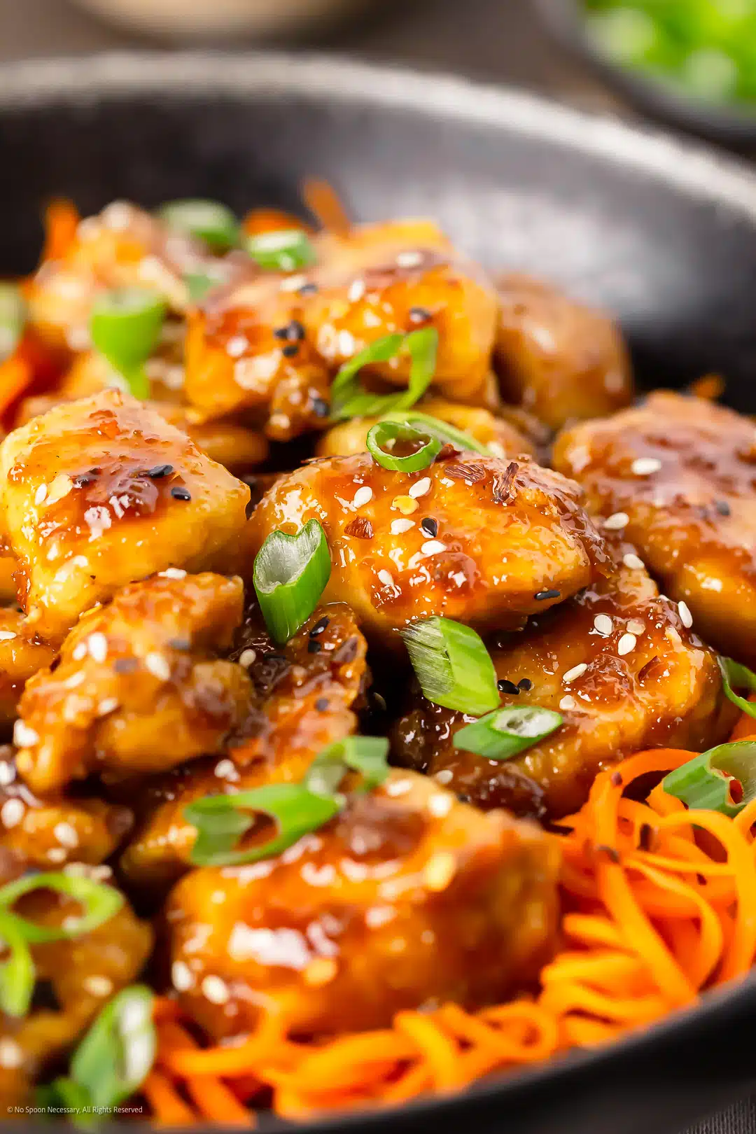 Close-up angled photo of a bowl of stir fry honey garlic chicken with spiralized carrots, sesame seeds, and green onions.