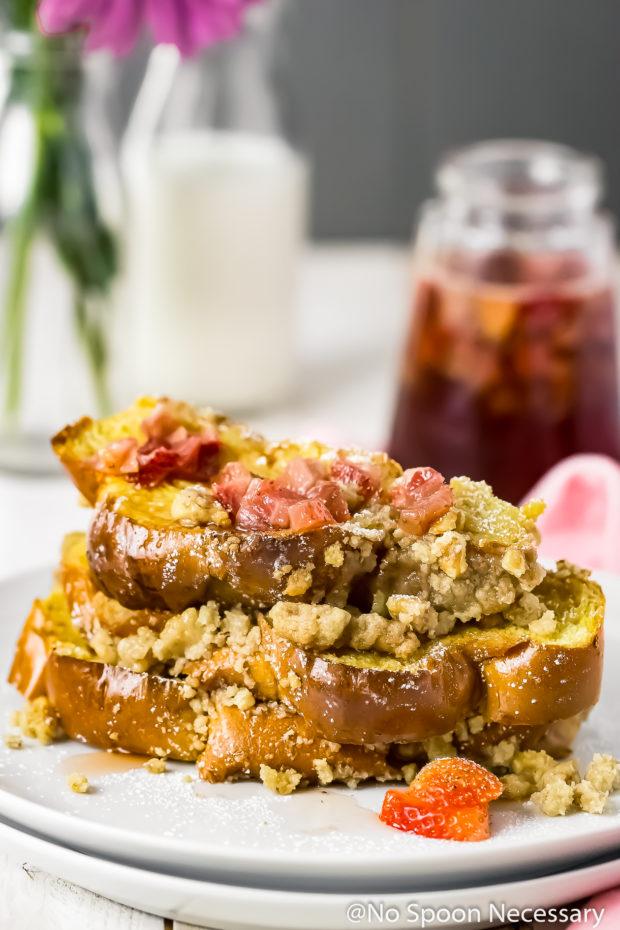 Straight on shot of a stack of Strawberry Baked Challah French Toast on a white plate with homemade strawberry maple syrup, small vase of pink flowers and a milk glass blurred in the background.