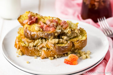 Straight on shot of a stack of Baked Strawberry French Toast on a white plate with homemade strawberry maple syrup and a jar of syrup, fork, knife and pink linen to the side of the plate.