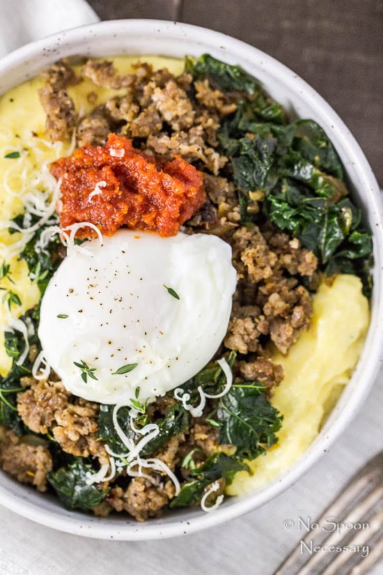Overhead, up-close shot of a Sausage Breakfast Polenta Bowls with kale, sausage, poached egg and sundried tomato pesto on a dark wood surface and neutral linen.