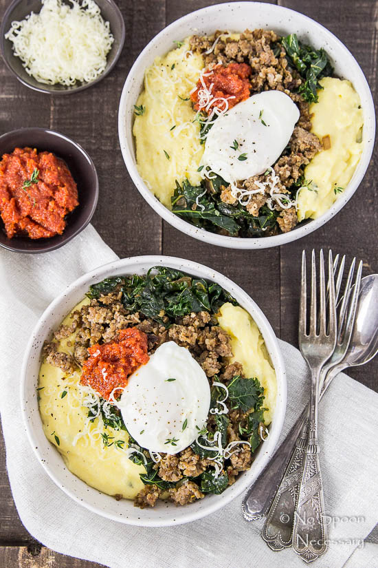 Overhead shot of two Sausage Breakfast Polenta Bowls with kale, sausage, poached egg and sundried tomato pesto on a dark wood surface and neutral linen with a stack of forks and spoons, and small bowls of pesto and shredded parmesan in the corner.
