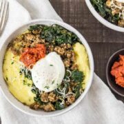 Overhead shot of two Sausage Breakfast Polenta Bowls with kale, sausage, poached egg and sundried tomato pesto on a dark wood surface and neutral linen with a stack of forks and small bowl of sun-dried tomato pesto arranged around the bowls.