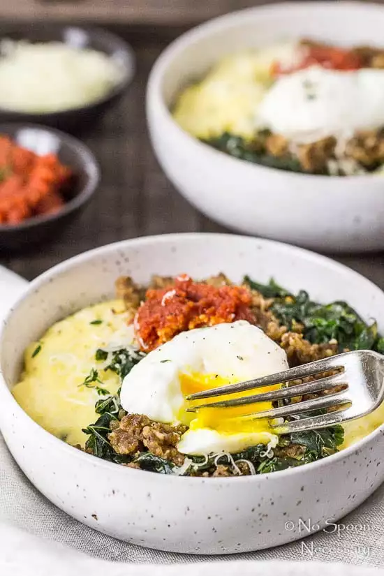 45 degree angle shot of a fork breaking the poached egg on a Sausage Breakfast Polenta Bowls with kale, sausage and sundried tomato pesto with another breakfast bowl, and small bowls of pesto and shredded cheese blurred in the background.