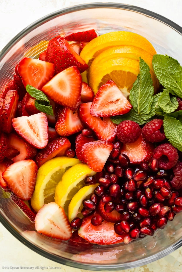Overhead photo of chopped and sliced fresh fruit - strawberries, raspberries, oranges and lemons - in a large glass serving bowl.