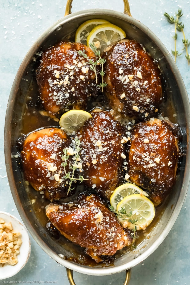 Overhead photo of Balsamic Fig Glazed Chicken garnished with lemon slices and fresh thyme in a round baking dish with a ramekin of chopped walnuts next to the dish.