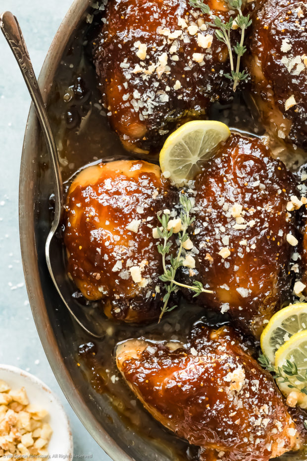 Overhead, up-close photo of Balsamic Fig Glazed Chicken garnished with lemon slices and fresh thyme in a round baking dish with a serving spoon inserted under one of the chicken thighs. 
