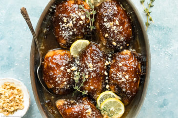 Overhead photo of Balsamic Fig Glazed Chicken garnished with lemon slices and fresh thyme in a round baking dish with a serving spoon resting under one of the chicken thighs and a ramekin of chopped walnuts next to the dish.