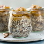 Straight on photo of three individual jars of Banana Overnight Oats on a neutral colored plate with spoons next to the jars and a glass jar of rolled oats blurred in the background.
