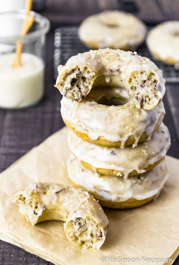 Straight on shot of a stack of Pecan Brown Sugar Baked Cake Donuts with Bourbon Maple Glaze, with one donut ripped in half, on a piece of folded brown parchment paper with a jar of glaze and additional donuts blurred in the background.