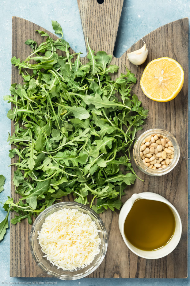 Overhead shot of all the ingredients needed to make Arugula Pesto neatly arranged on a gray wood serving board.