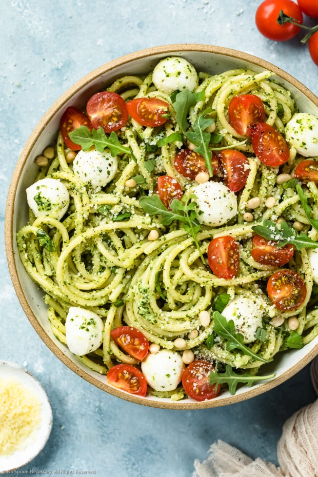 Overhead shot of Arugula Pesto Pasta tossed with mozzarella pearls and halved cherry tomatoes in a white bowl with a ramekin of grated parmesan cheese and vine-ripe cherry tomatoes arranged around the bowl.