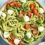 Overhead shot of Arugula Pesto Pasta tossed with mozzarella pearls and halved cherry tomatoes in a white bowl with a ramekin of grated parmesan cheese, a tan linen and vine-ripe cherry tomatoes arranged around the bowl.
