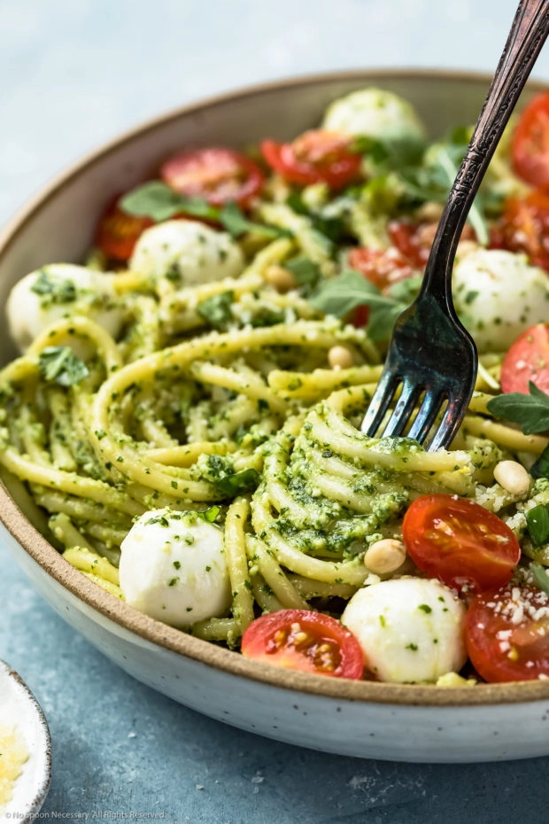Angled, up-close shot of Arugula Pesto Pasta in a white bowl with a fork inserted and twirled around the pasta to showcase the pesto.