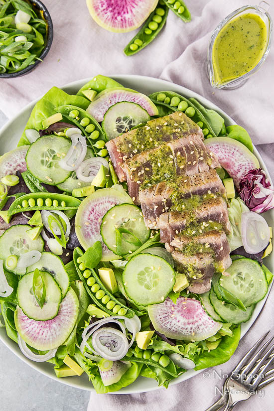 Overhead shot of an Ahi Tuna Spring Salad on a white plate with a pale purple linen under the plate and a small ramekin of sliced scallions and Asian Vinaigrette in a small glass creamer in the top corners of the shot. 