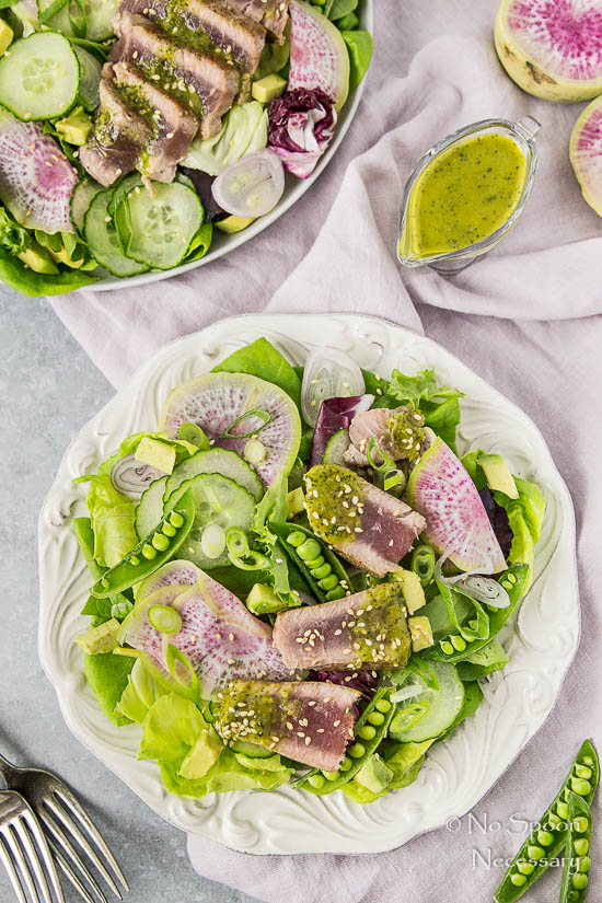 Overhead shot of an Ahi Tuna Spring Salad on a white plate with a pale purple linen under the plate and an additional salad, small glass creamer of Asian Vinaigrette and forks surrounding the main plate.