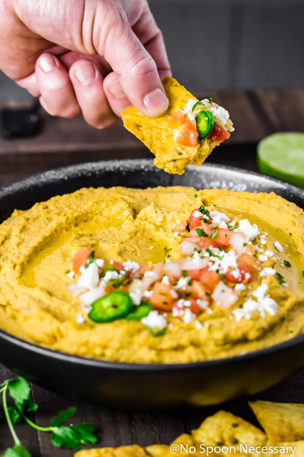 Angled shot of a black bowl filled with Fajita Hummus and a hand holding a pita chip dipping it into the hummus.